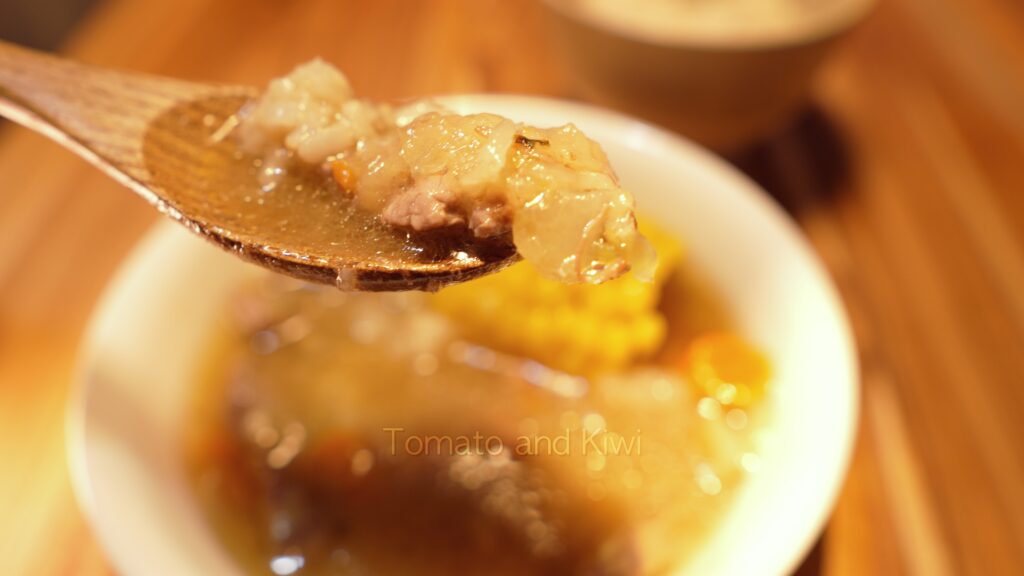 A wooden spoon filled with rich Instant Pot Beef Nilaga broth. A piece of meat dangles just off the spoon. The background has a blurred view of the bowl of Beef Nilaga.